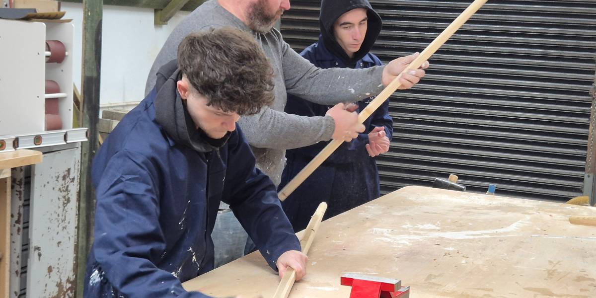 3 People in a workshop environment. 2 are wearing work suits while the third is wearing a jumper and jeans. He is showing one of them how to sand a bit of wood.