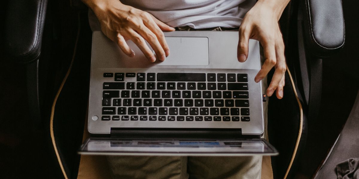 A laptop sits on a persons lap as they rest in a chair. It is a top down view with a pair of hands coming into the frame and using the mouse pad.