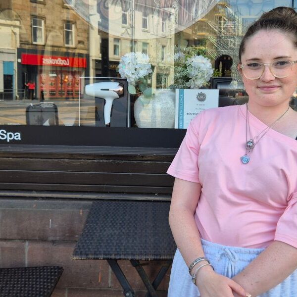 Girl standing in front a store front, smiling. Table and chair can be seen in outside the store.
