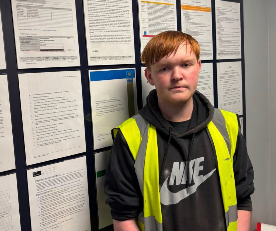 Young Man in a Hi Vis stop standing in font of a wall with various pieces of paper stuck to it.